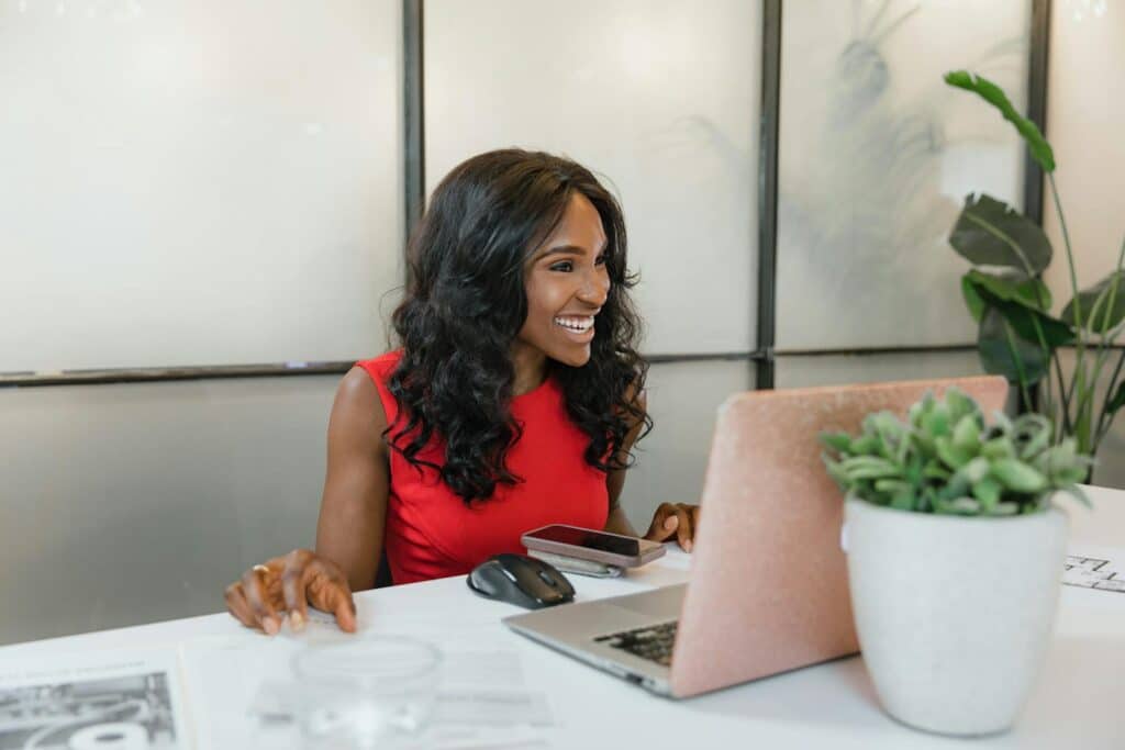 Professional black woman smiling at desk using laptop and smartphone in office.